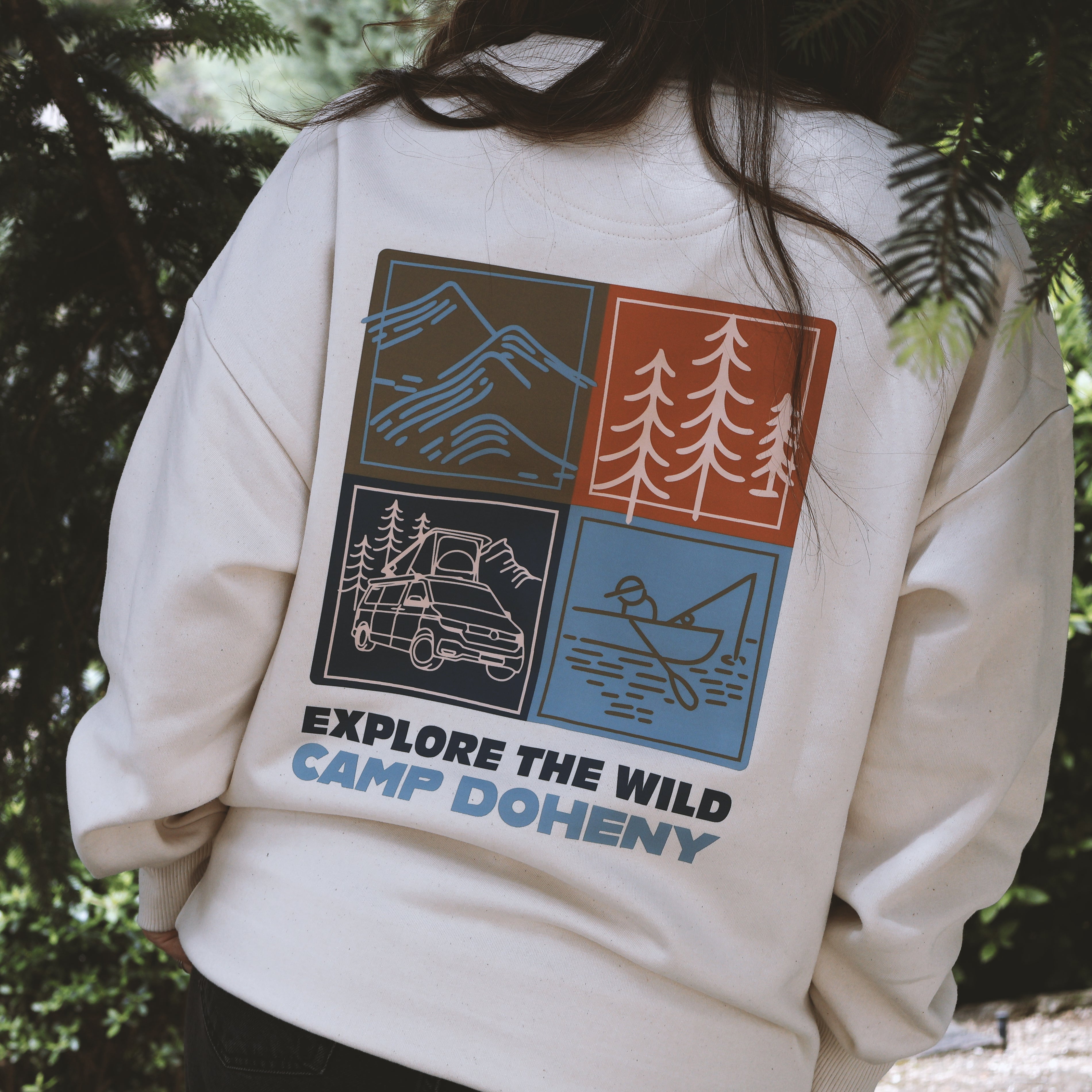 Beige sweatshirt with outdoor-themed graphics and text 'Explore the Wild Camp Doheny' on a person against a forest background.